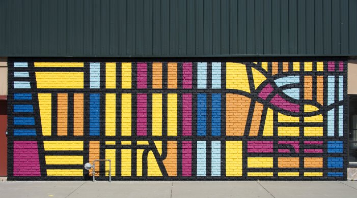 A brick wall patterned with bright geometric forms in shades of magenta, sky blue, navy, yellow, and orange and thickly outlined in black fills much of the center of this image; a concrete sidewalk is visible along the bottom edge.
